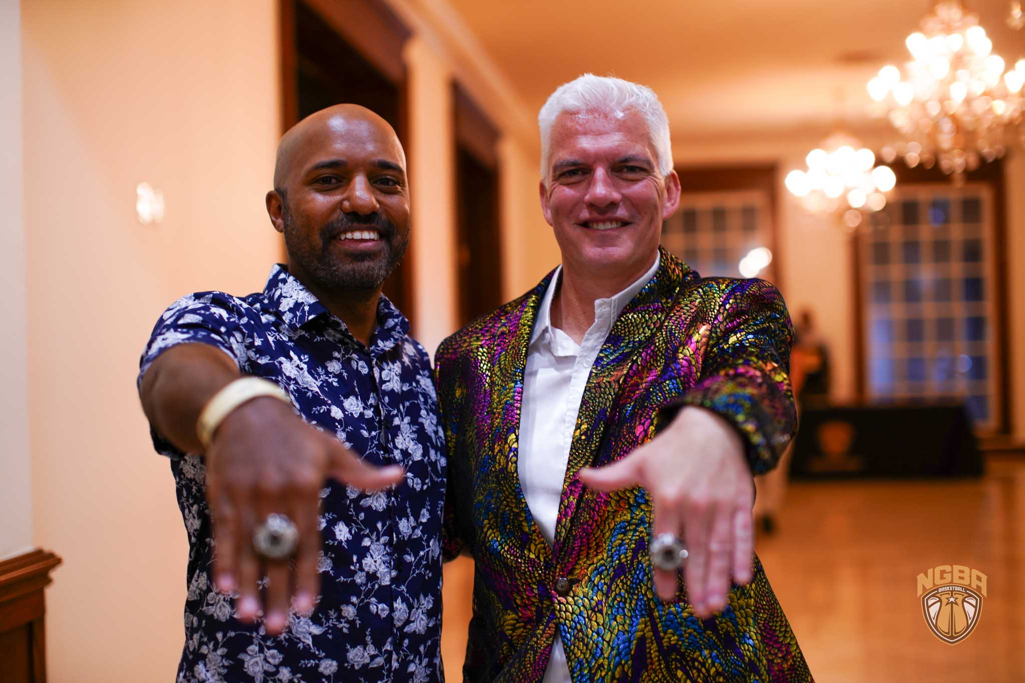 Two men smile for a photo while holding out their hands showcasing rings